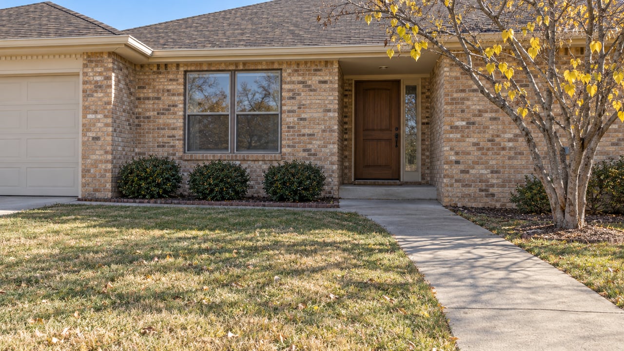 Low angled view across an autumn lawn and concrete approach toward a recessed wood front door in a buff-brick single-story home.