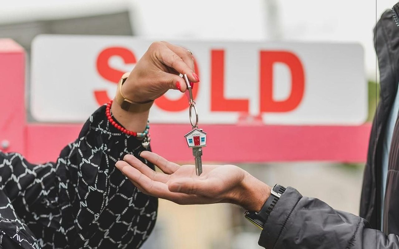 Two people exchange a house key in front of a SOLD sign.