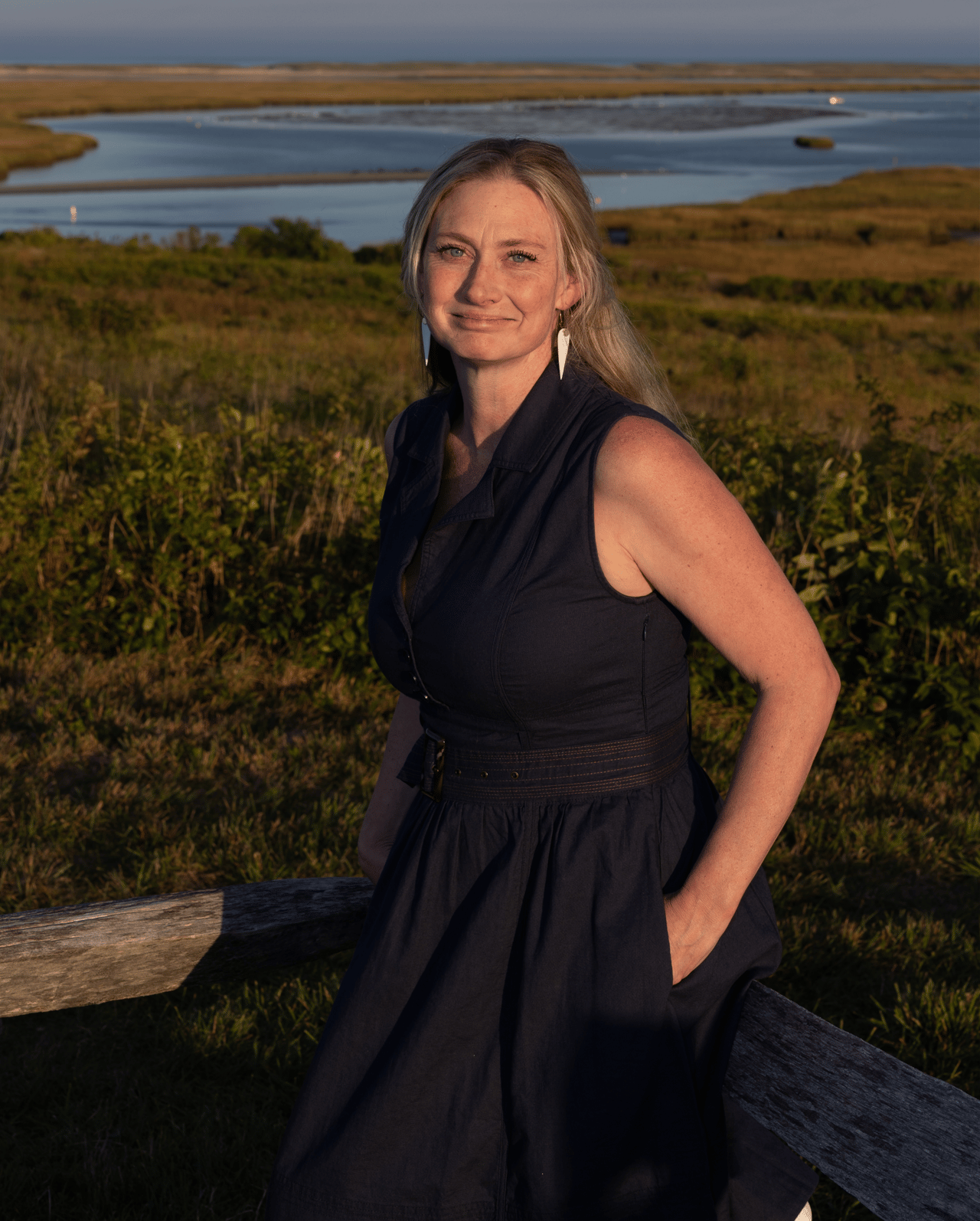 Shannon Burke in a dark dress beside a marsh inlet at golden hour.