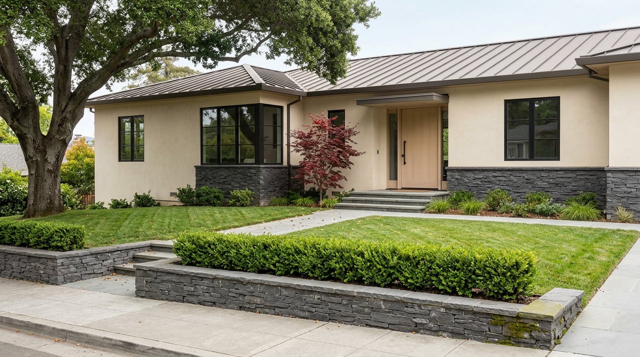 Hillside home with ivory stucco, charcoal stone retaining wall, terraced lawn, and a mature oak shading the roof.