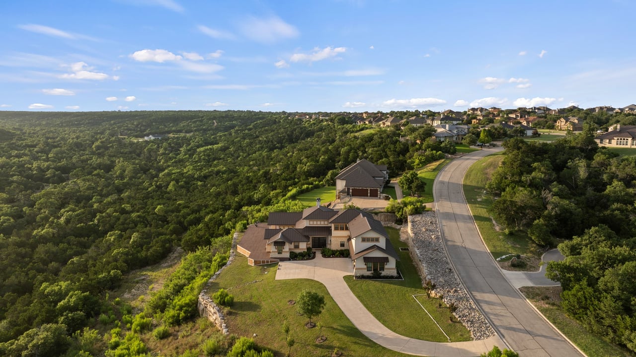 Large home beside a wooded hillside, viewed from above with a curving road nearby.