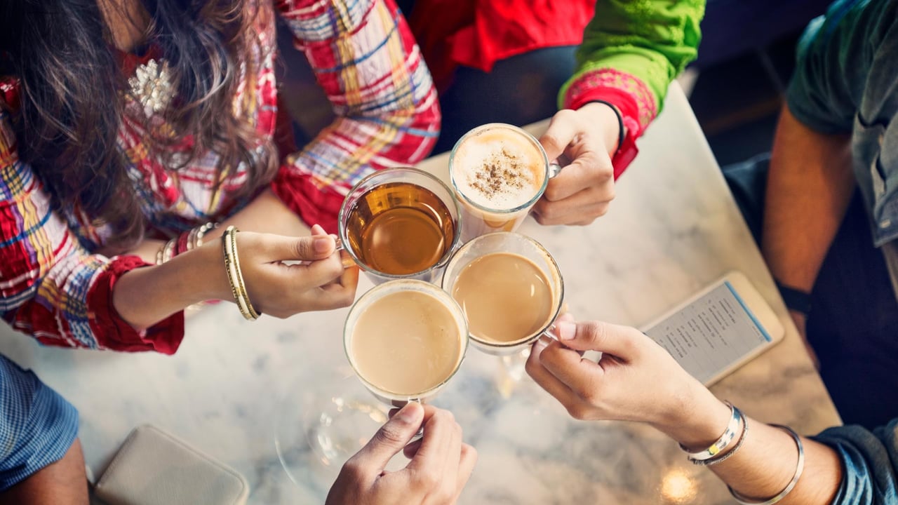 A top-down view of millennial friends making a toast with clear glass mugs of coffee, espresso lattes, and tea at a café table, illustrating the vibrant local café culture and community lifestyle enjoyed by young professionals living in Burlington Ontario, Oakville, and Mississauga.