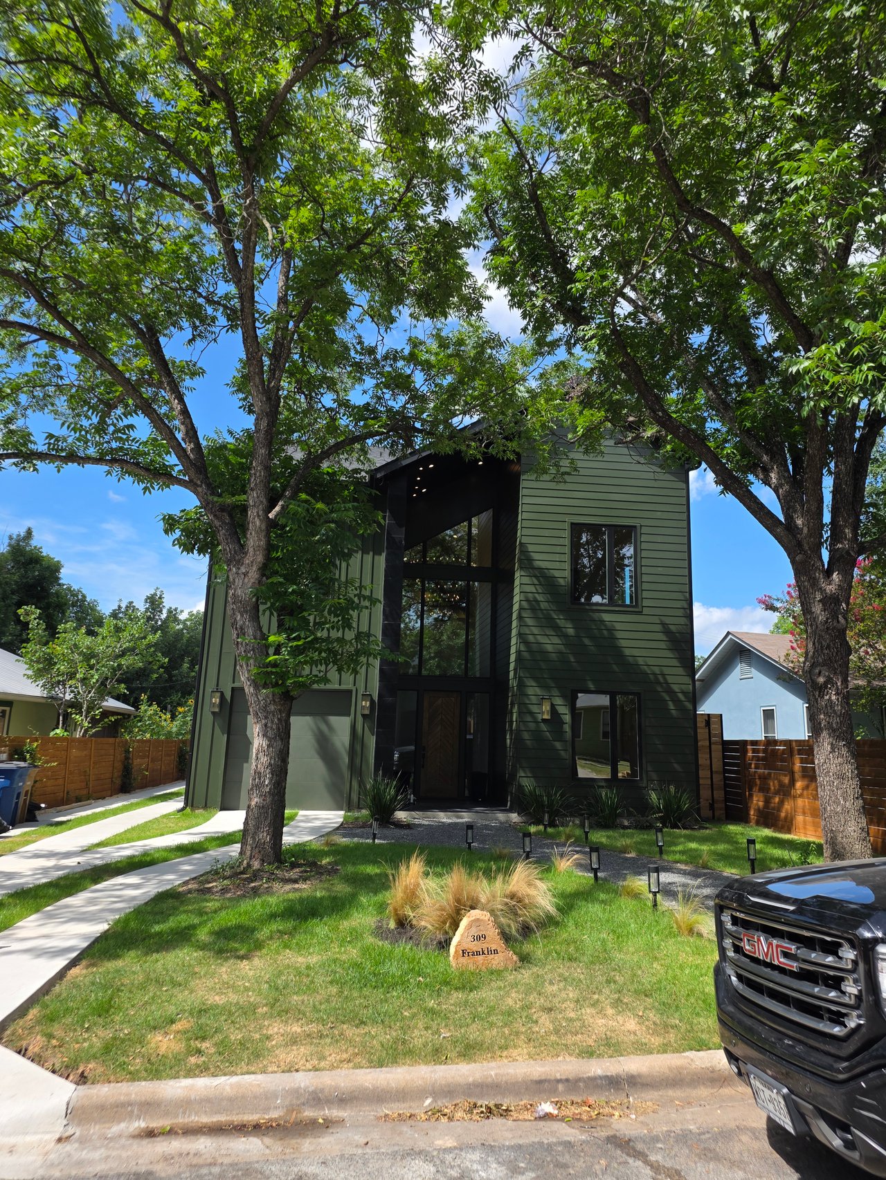 Modern green two-story home framed by mature trees, with a tall glass entry and front lawn.
