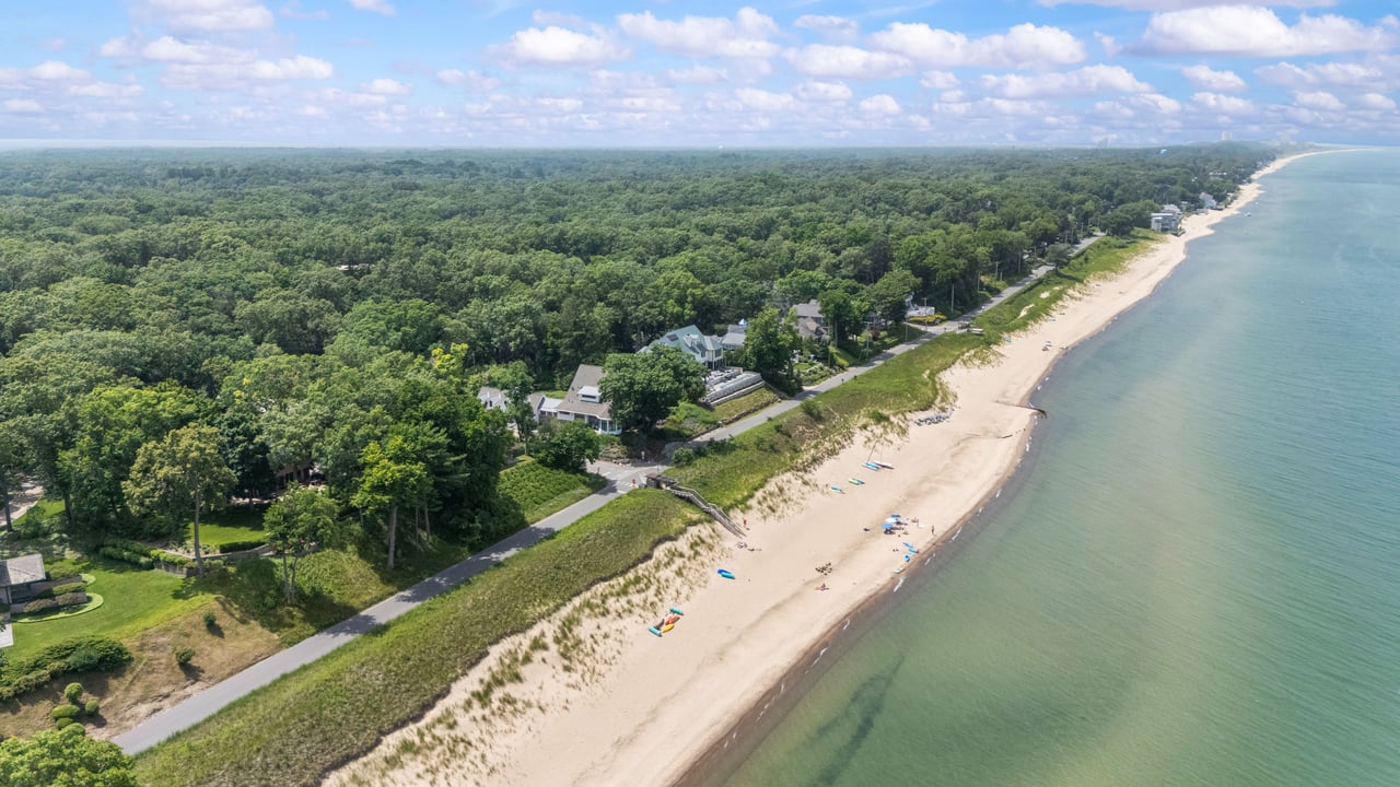 Aerial view of a sandy shoreline, wooded homes, and a coastal road beside calm water.