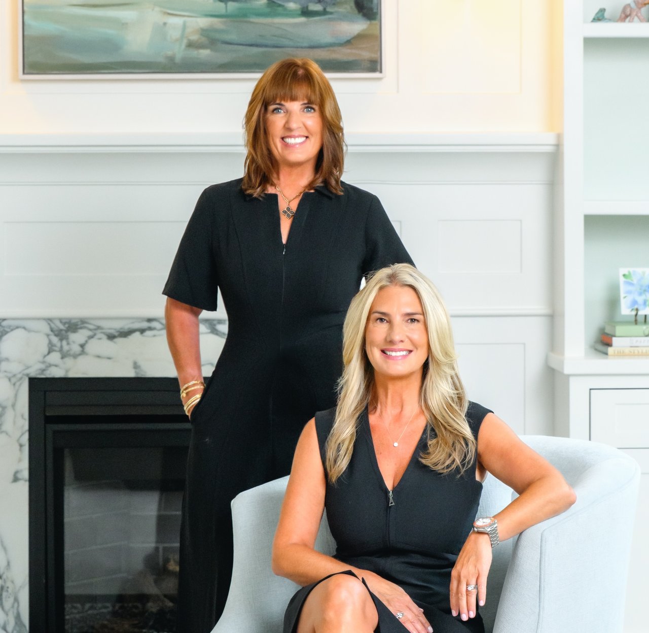 Two women pose together, one standing behind a seated woman beside a marble fireplace.