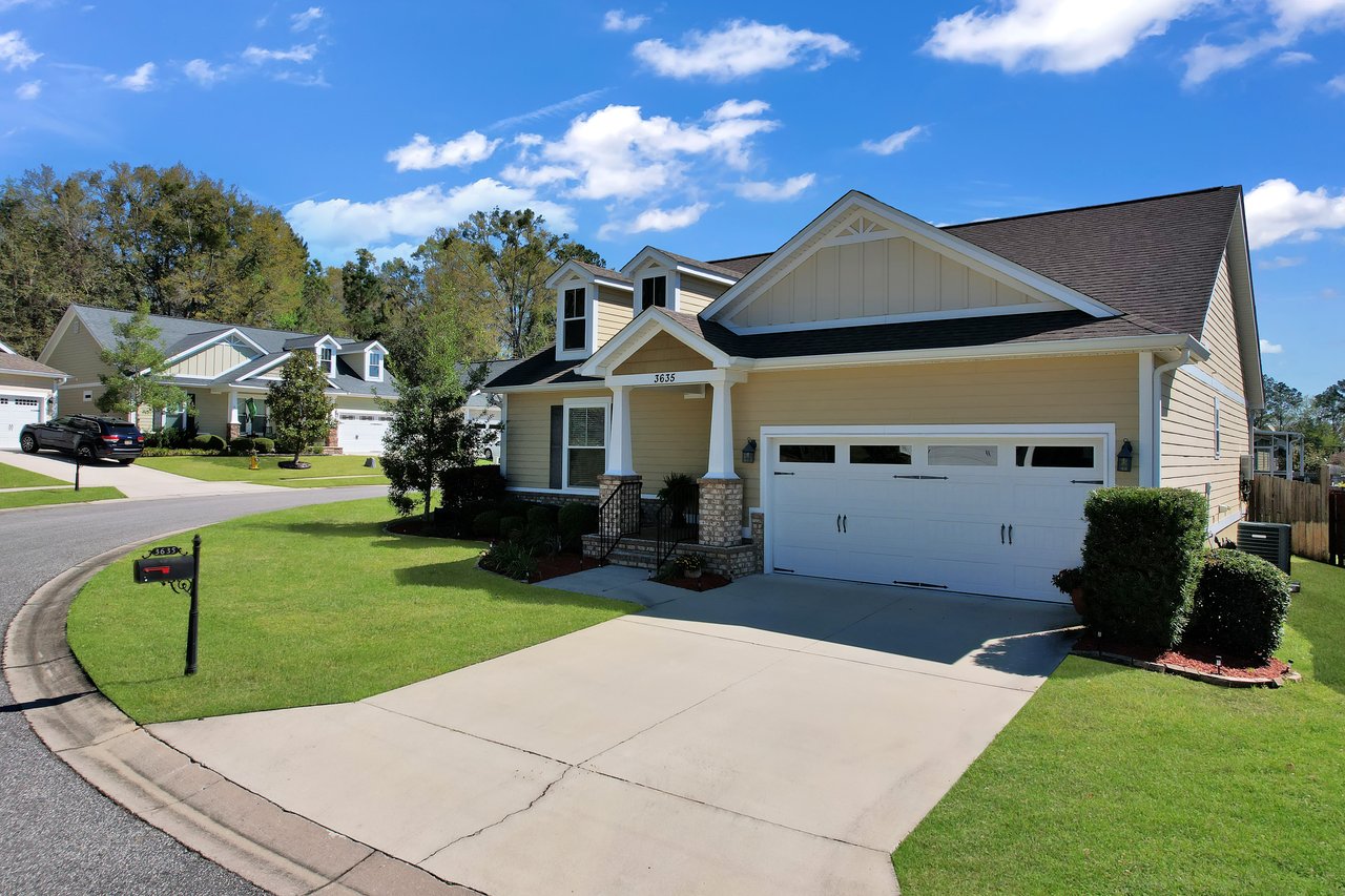 Suburban house with beige siding, white trim, and a two-car garage. Manicured lawn and shrubs line the driveway. Bright, blue sky with fluffy clouds.