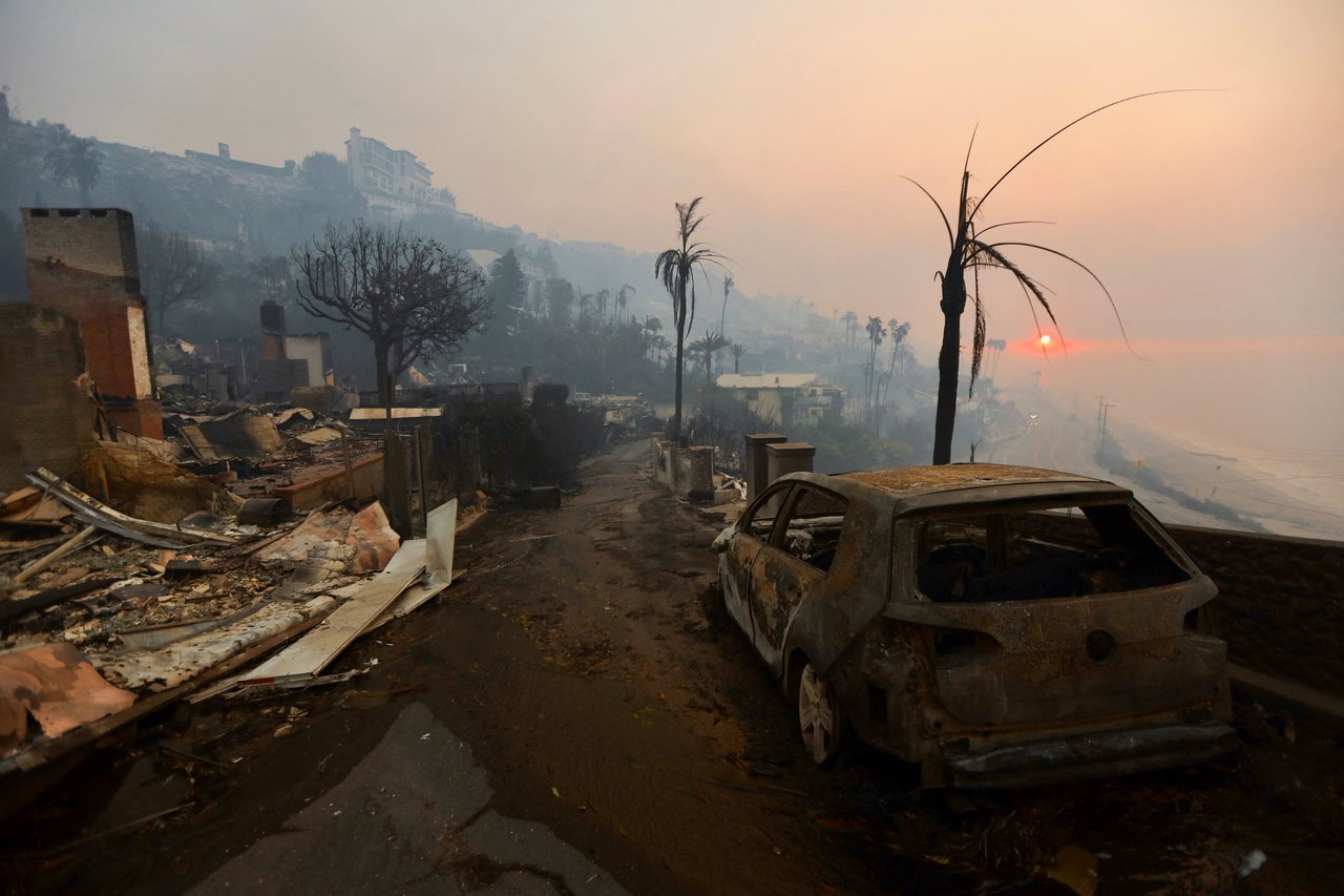  A dark, smoky landscape shows the aftermath of a fire, with charred remnants of houses and trees under a hazy, orange sky.