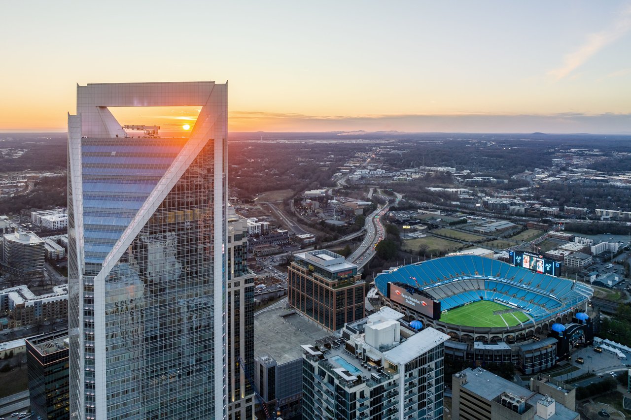 Aerial city skyline at sunset with a glass tower and blue-seated football stadium.
