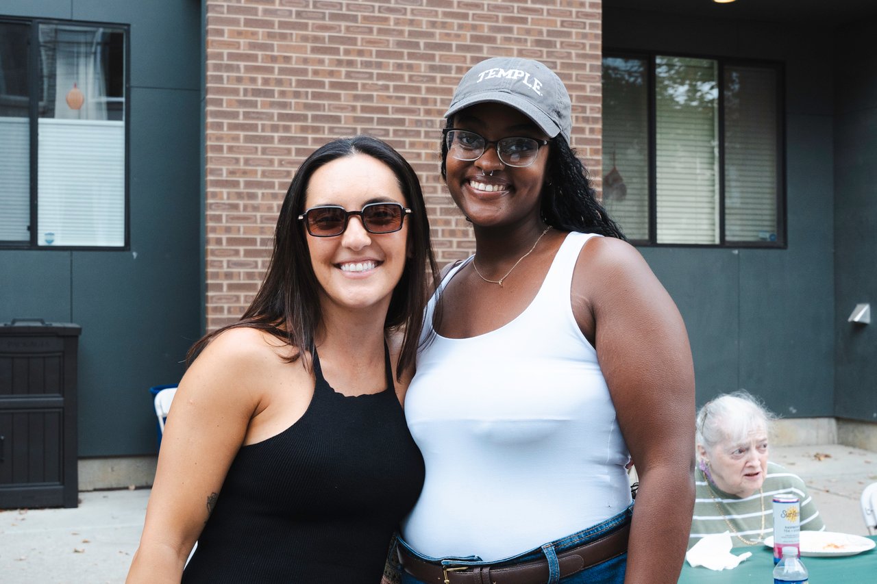 Two women pose together at a block party, with an older woman seated at a table behind them.