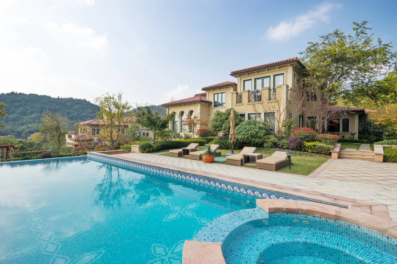 Mediterranean-style home behind a swimming pool and curved spa, with landscaped grounds and wooded hills beyond.