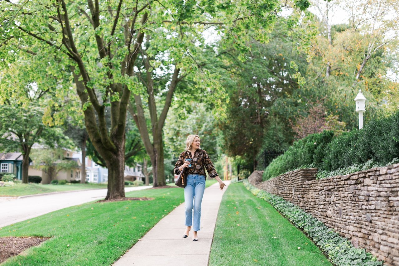 A woman holding a coffee walks along a tree-lined residential sidewalk in Madison, Wisconsin, beside a stone retaining wall.