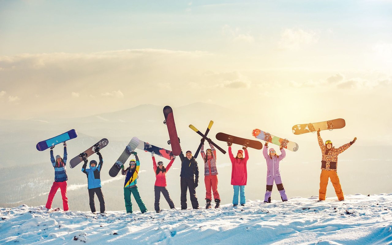 Snowboarders in colorful winter gear raise their boards on a snowy mountain ridge.