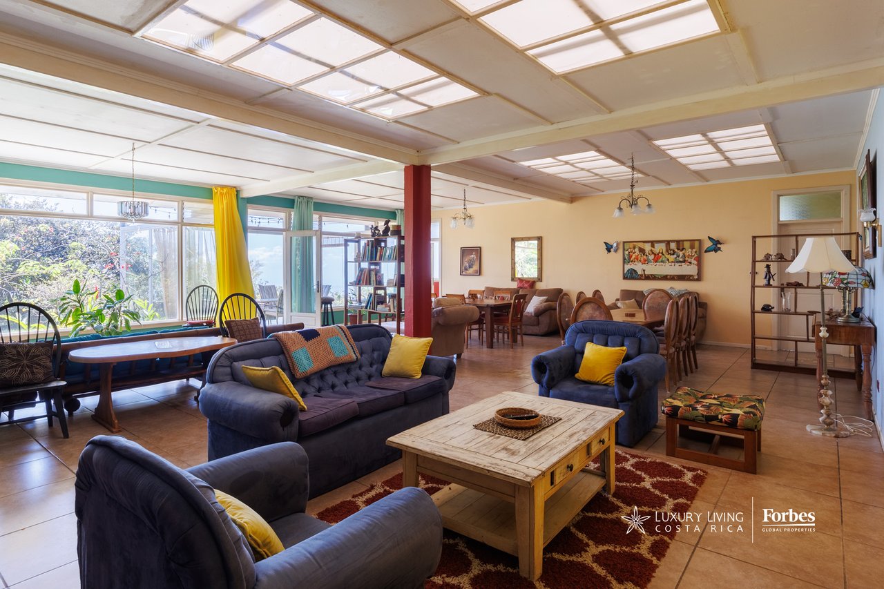 Open-plan living room with blue sofas, dining areas, and large windows overlooking greenery.