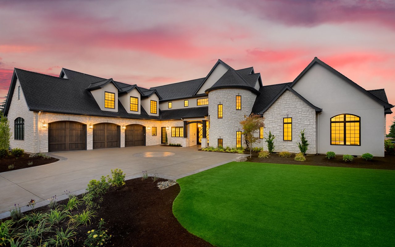 Large stone house with a circular turret, multiple garage doors, and a broad lawn at dusk.