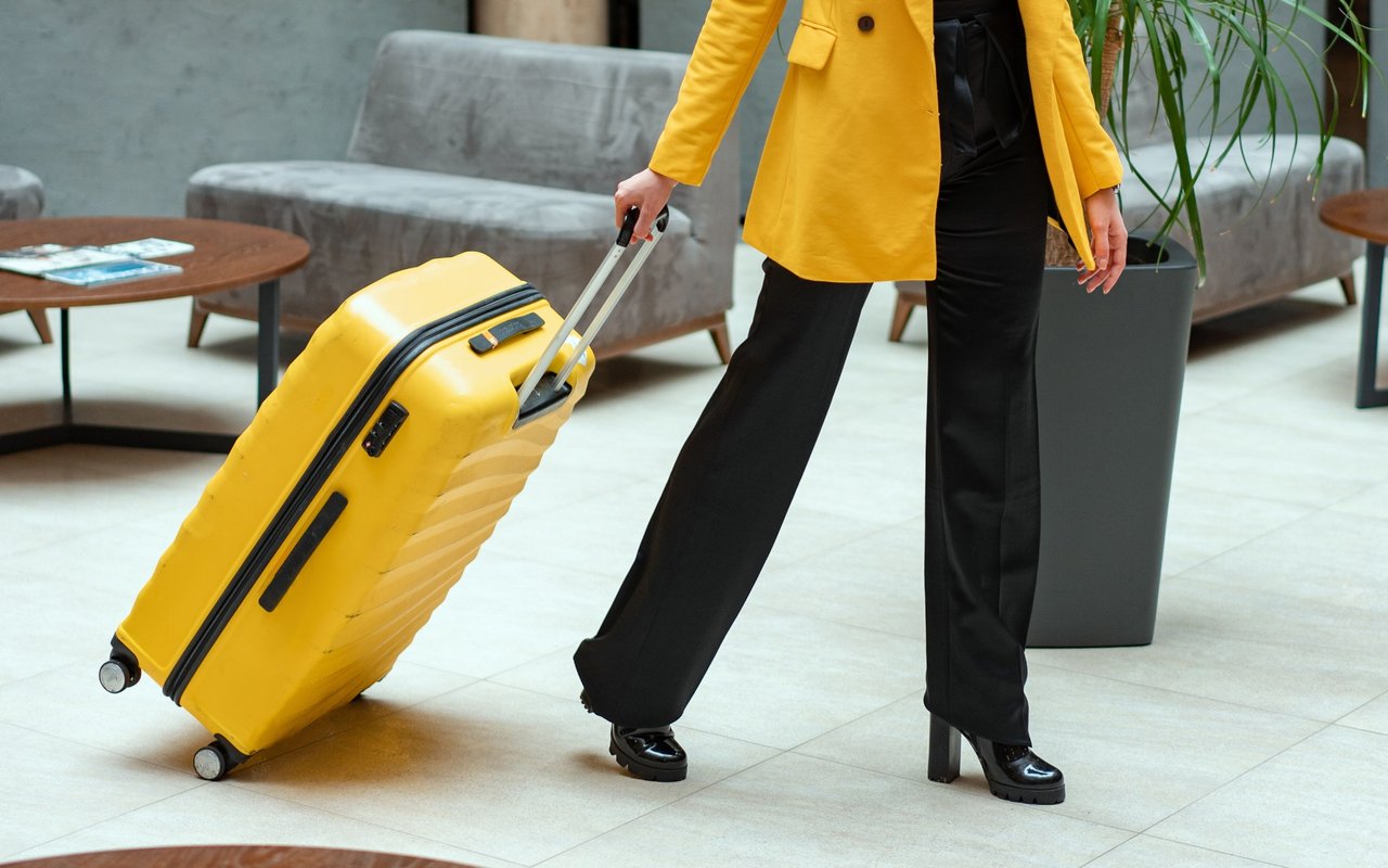 Traveler in a yellow coat pulling a matching rolling suitcase through a modern lobby.