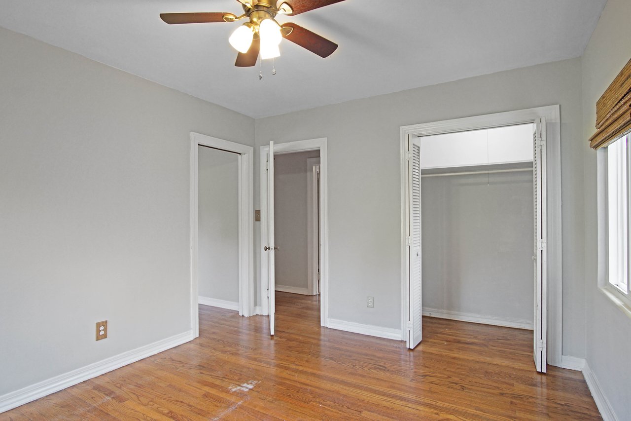 Empty room with light gray walls, wooden floor, a ceiling fan with lights, open closet doors, and windows with wooden blinds. Bright and neutral.