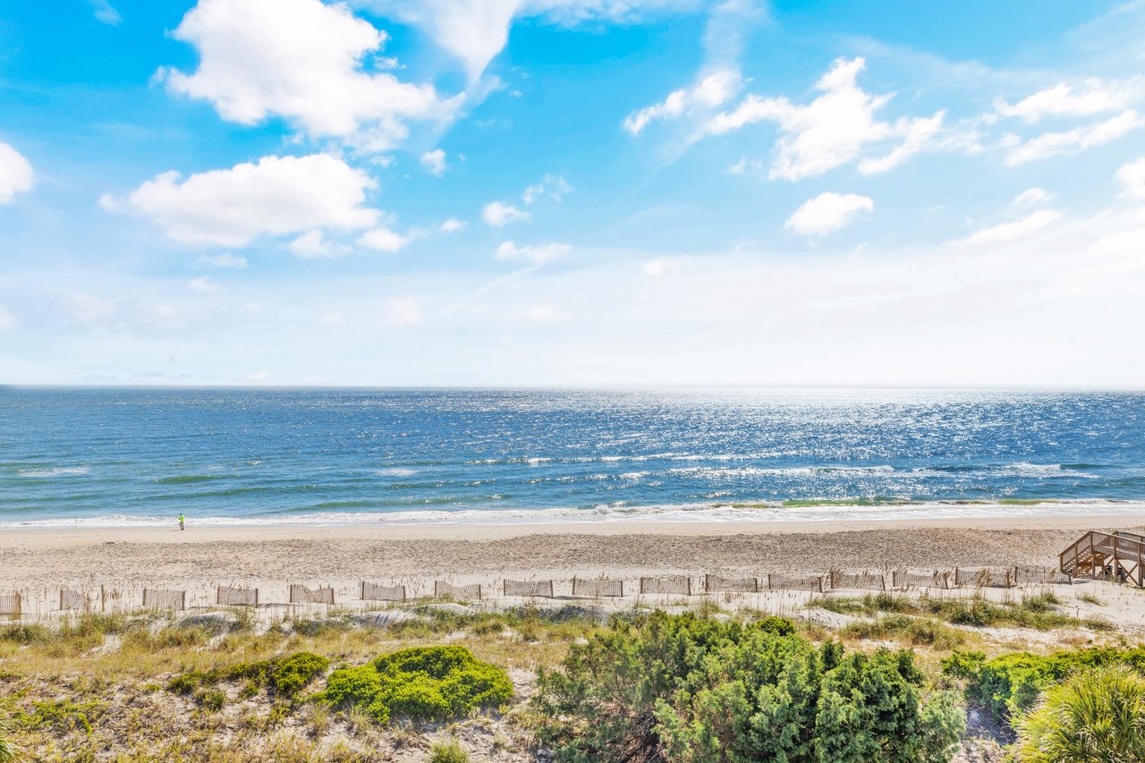Sandy beach and ocean horizon with dune vegetation and a wooden stairway beneath a bright, cloudy sky.