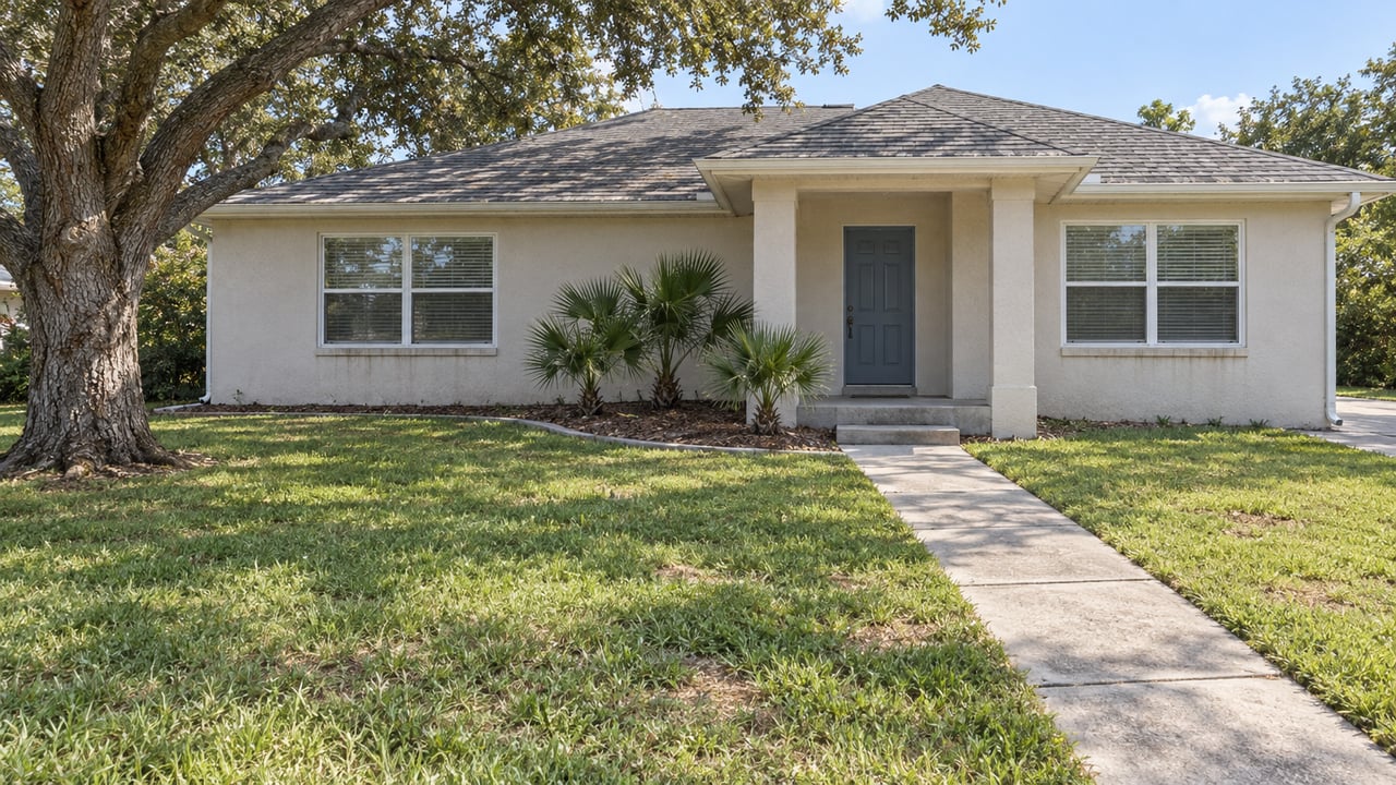 A single-story off-white ranch home behind a deep lawn, with a curved concrete walk, a mature live oak, and dwarf palmettos beside the covered entry.