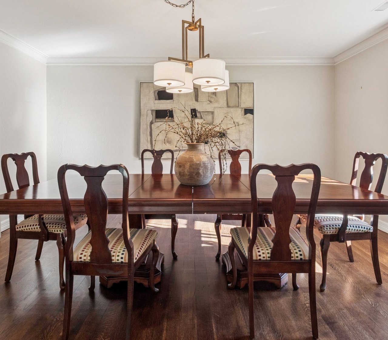 Furnished dining room with a dark wood table, carved chairs, brass chandelier, and vase of dried branches.