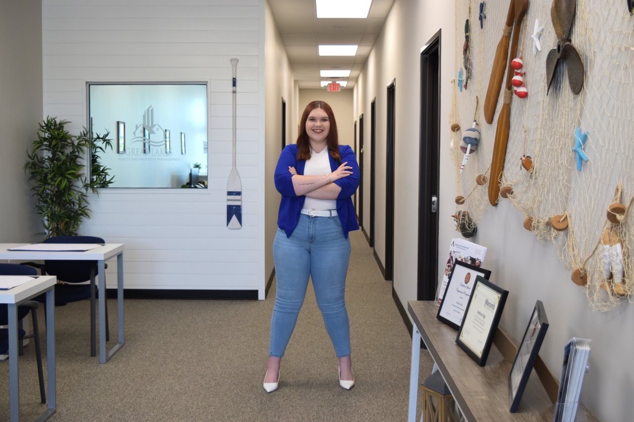 Real Estate Agent, Megan, standing with her arms crossed in the Great Lakes Realty & Property Management office in a blue blazer with her arms crossed while smiling at the camera