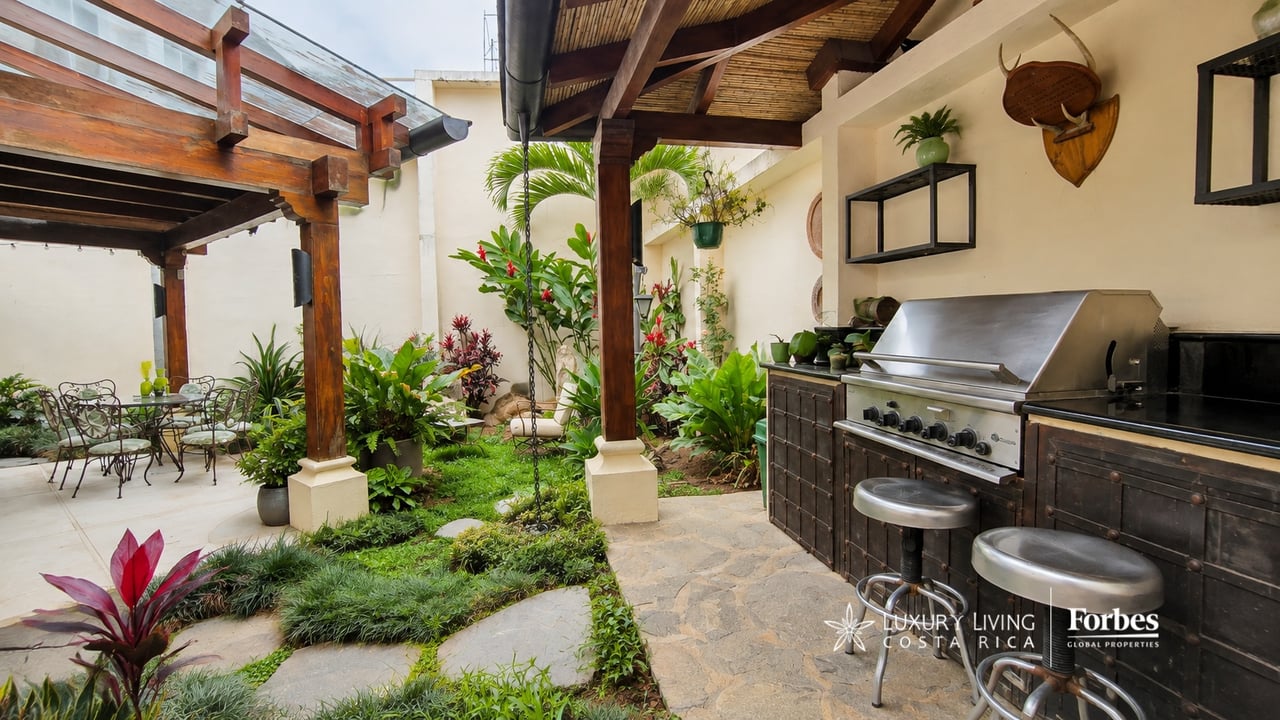 Covered outdoor kitchen with stainless-steel grill beside a lush garden and timber pergola.
