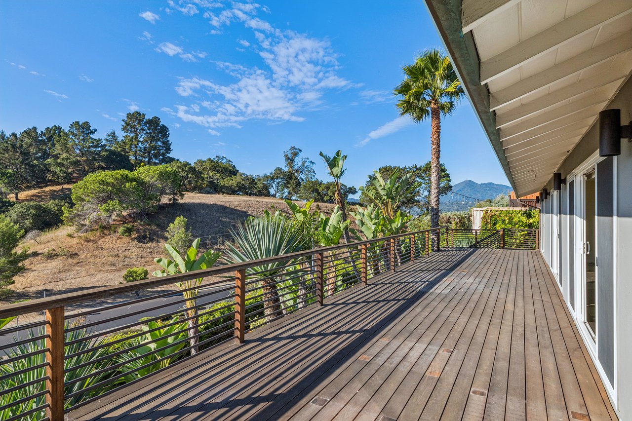 Wide wooden deck with dark railings, tropical plants, and distant mountain views.
