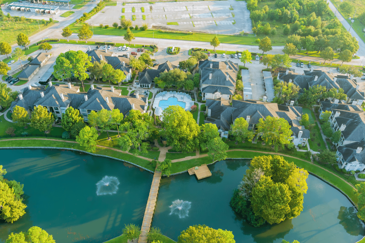 Aerial view of a Dallas apartment community featuring a central lake, walking paths, mature trees, and resort-style amenities surrounded by residential buildings.