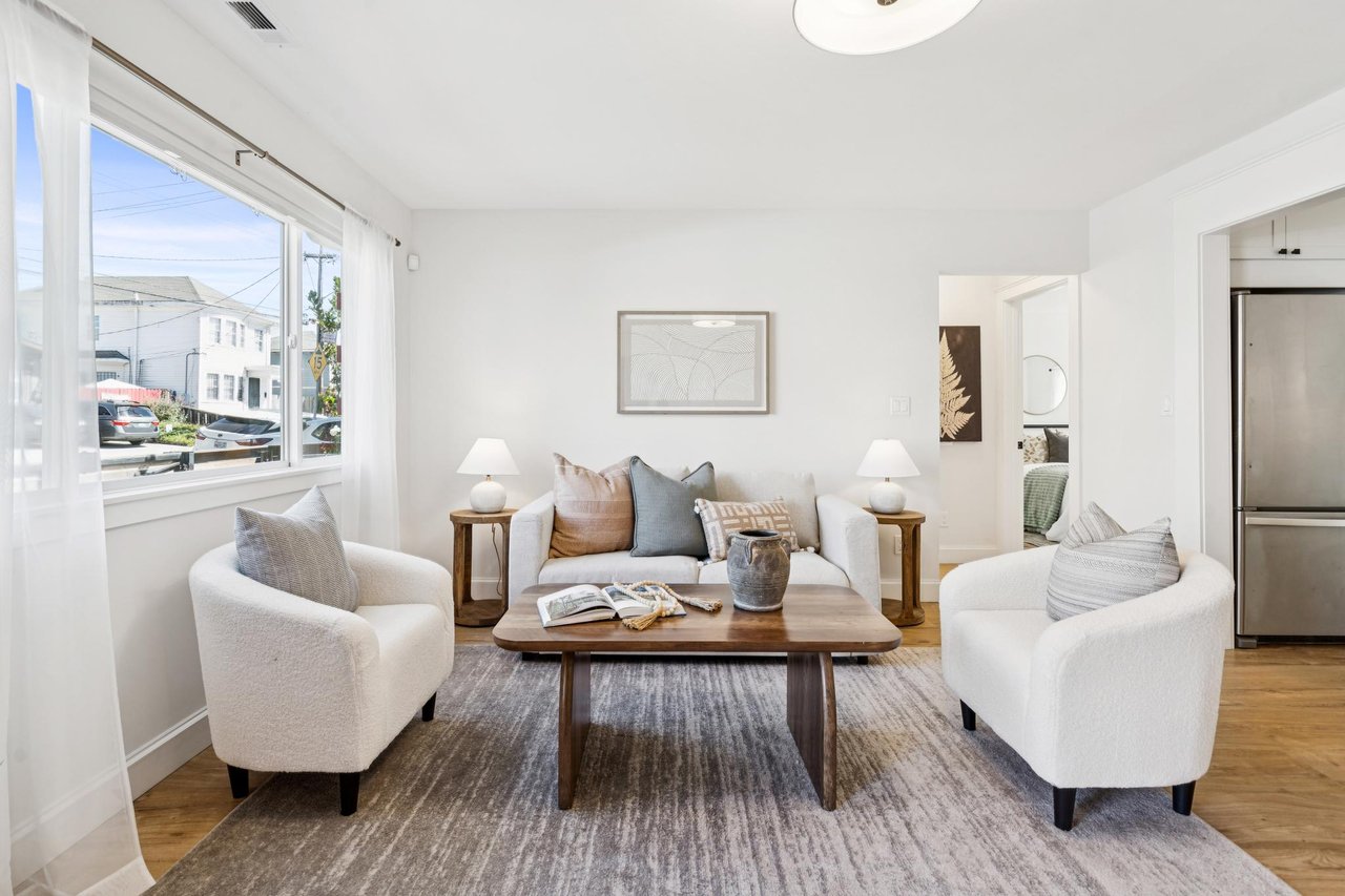 Staged living room with white armchairs, a wooden coffee table, and large street-facing windows.