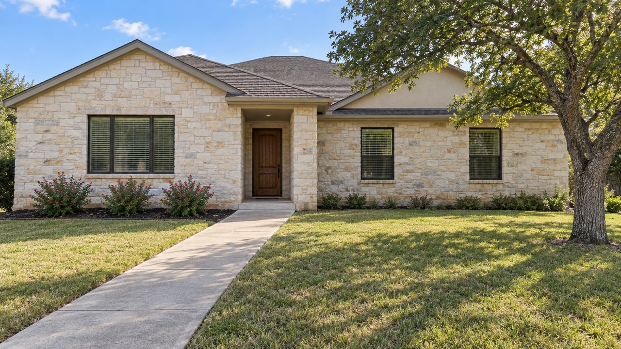 A single-story pale stone home with dark gabled roof, recessed oak entry, broad autumn lawn and one mature tree.