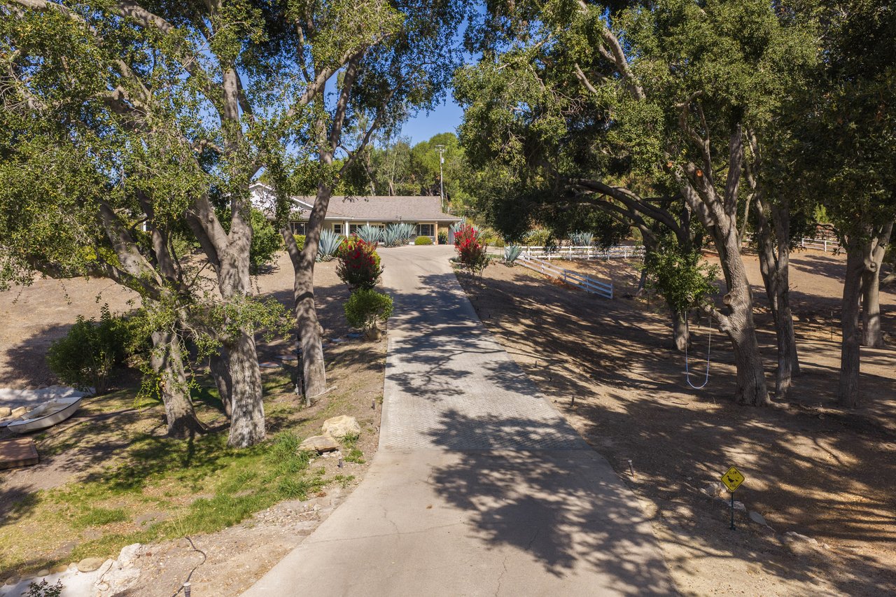Single-story home at the end of a tree-lined driveway, with agave plants and a white fence.
