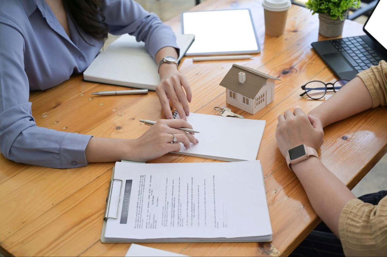 Two people review paperwork beside a model house and keys at a wooden table.