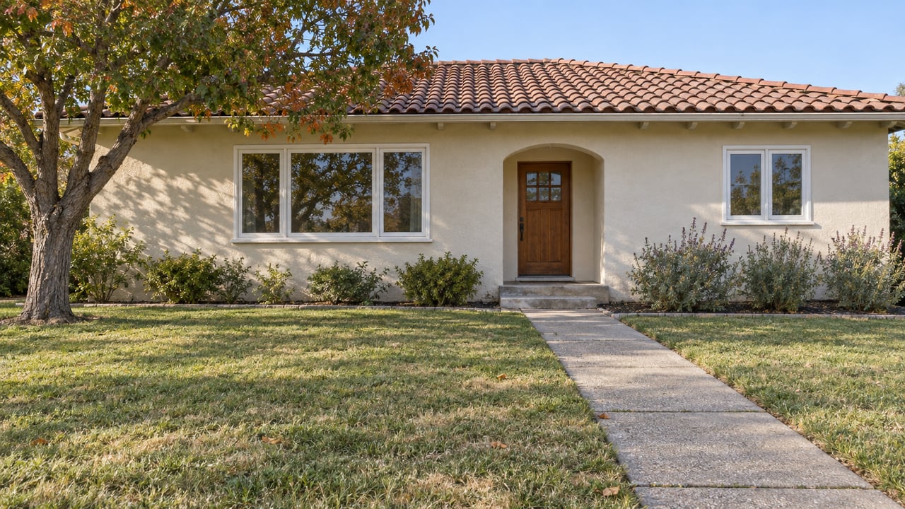 A single-story ivory stucco home with a clay tile roof, arched entry and deep front lawn in morning sunlight.