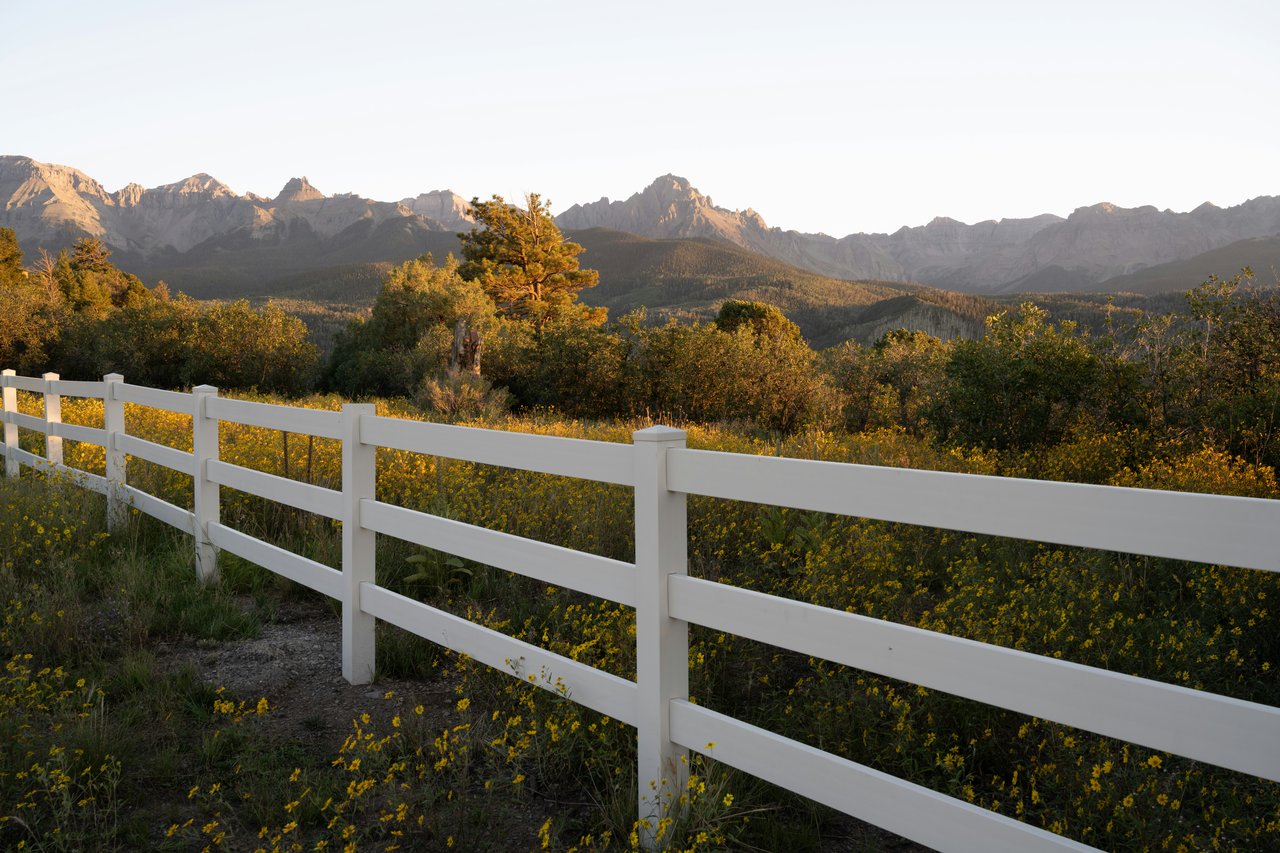 Panoramic mountain range beyond autumn-colored shrubs and a white ranch fence in warm evening light.