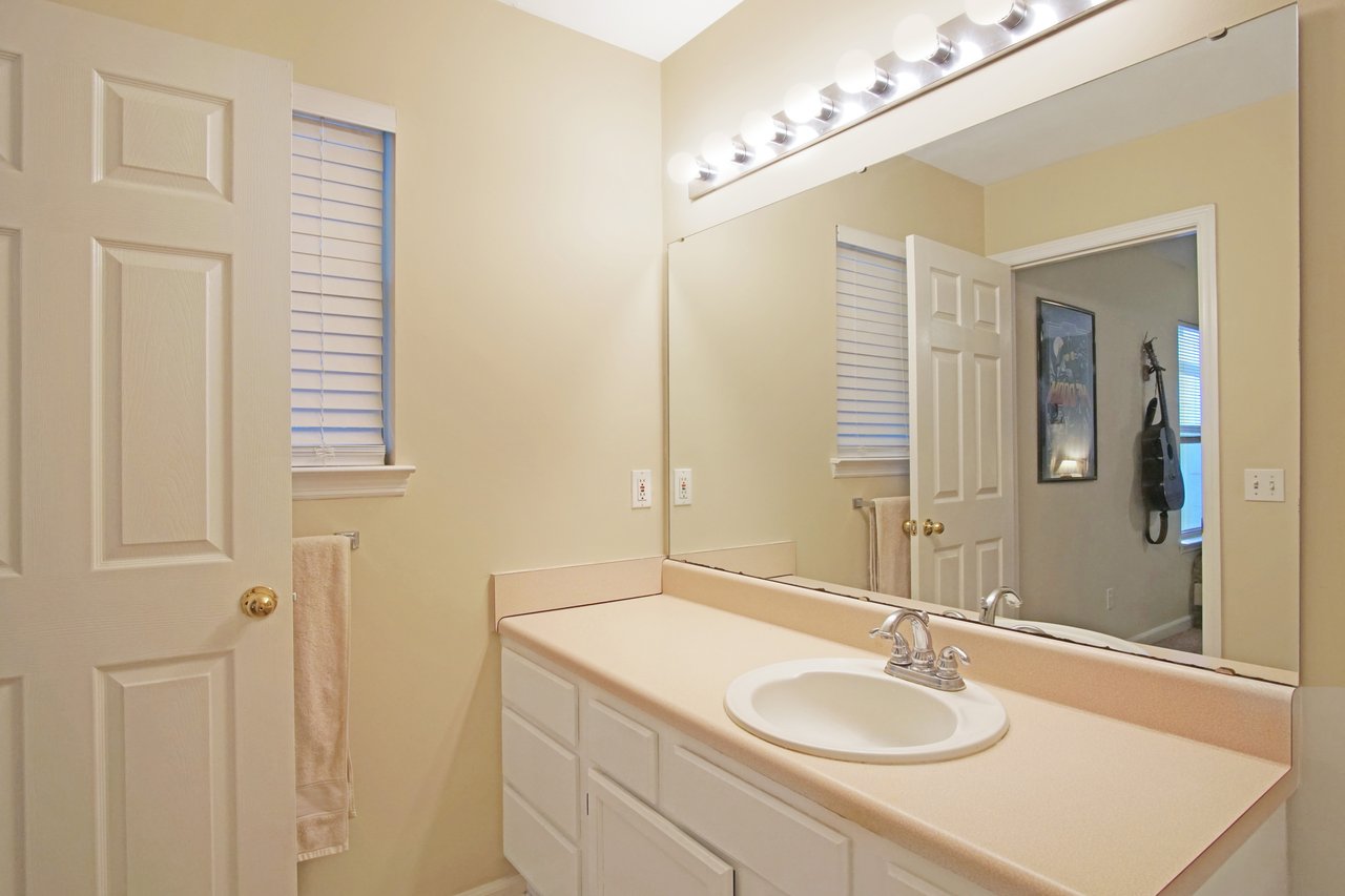 Bathroom at 529 Collinsford Road, Tallahassee, Florida, featuring a single-sink vanity with an extended countertop, large wall mirror with bright vanity lighting, and beige walls. The space includes white cabinetry, a towel rack, and natural light from two small windows with blinds.