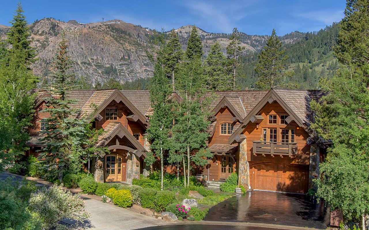 Wood-and-stone mountain home with peaked roofs, landscaped entrance, and forested mountain backdrop.
