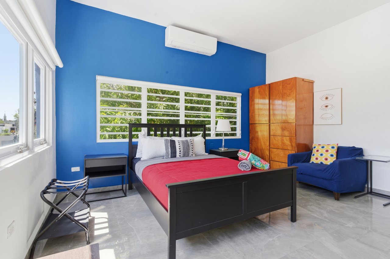 Furnished bedroom with a black bed, vivid blue accent wall, large windows, and blue armchair.