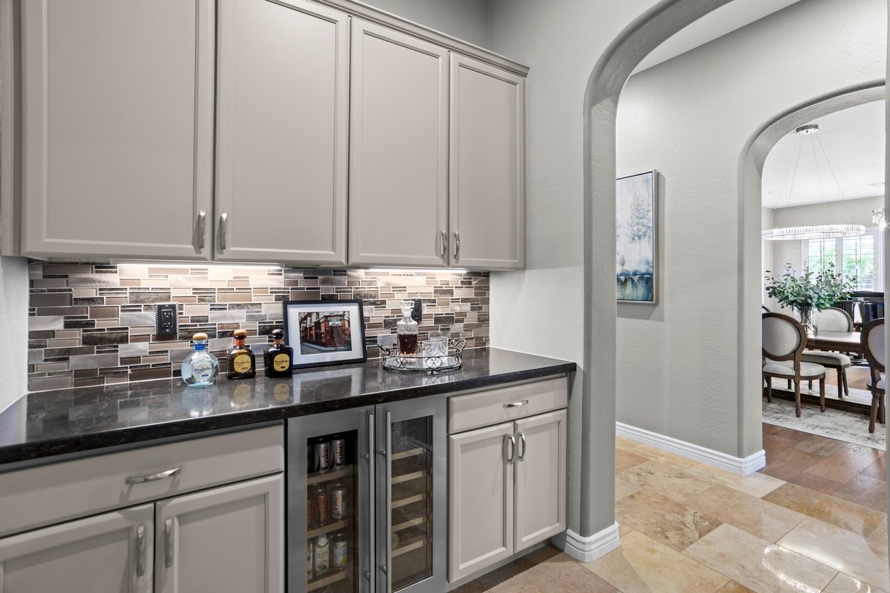 Built-in bar cabinet with upper and lower storage, mosaic tile backsplash, dark countertop, and a small beverage fridge, opening to a dining room through an arched hallway.
