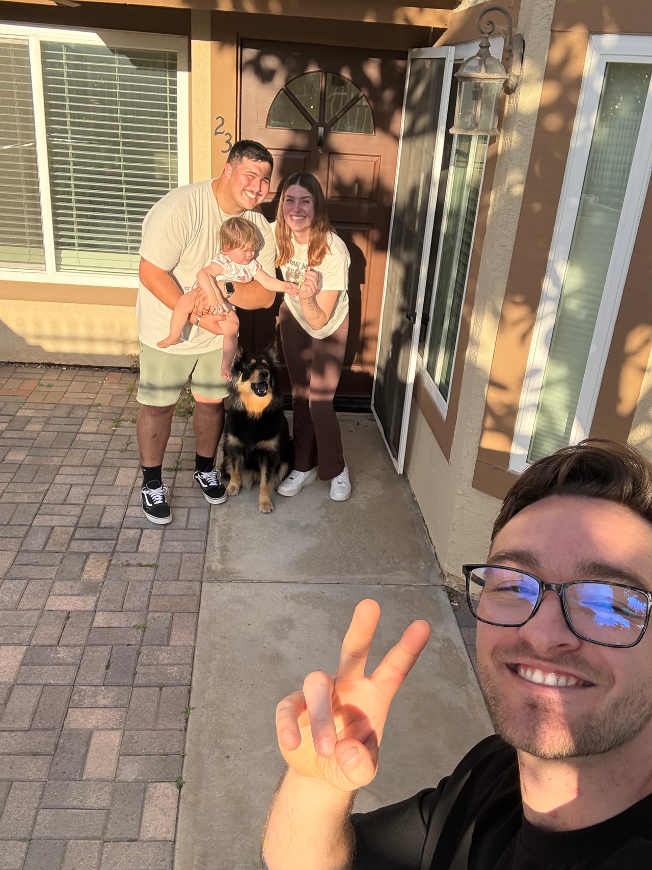 A man takes a selfie with a couple, baby, and dog outside a home.