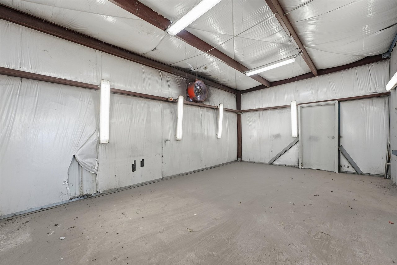 Empty garage with insulated metal walls, exposed beams, fluorescent lights, and a torn wall covering.