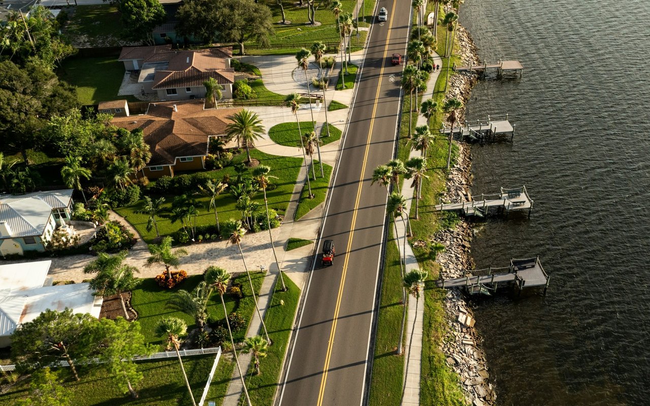 Waterfront homes and private docks line a palm-lined road beside the water.