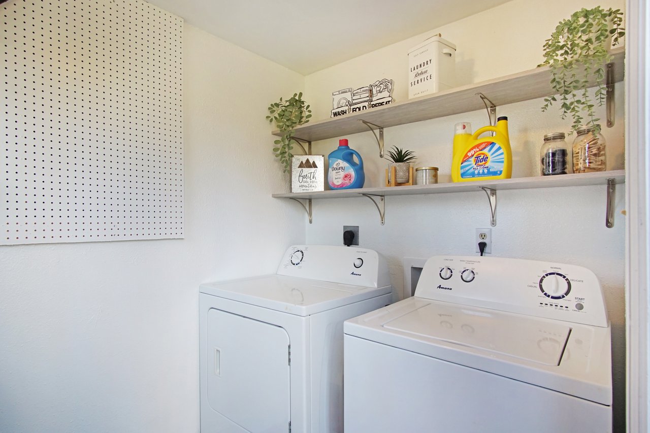 A tidy laundry room with a washer and dryer below a wooden shelf holding detergent, plants, and storage jars. A pegboard is on the left wall.