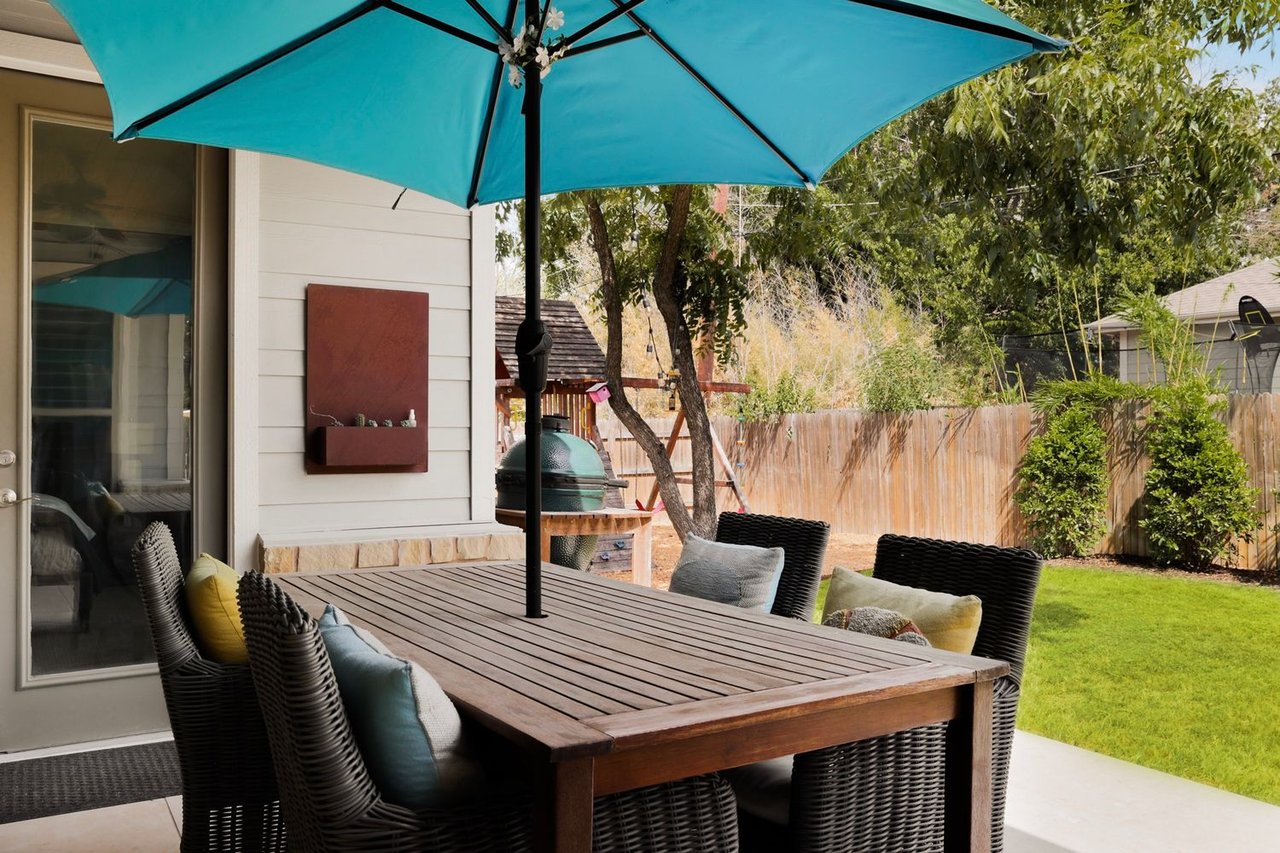 Patio dining set beneath a turquoise umbrella, with a ceramic grill and fenced backyard.
