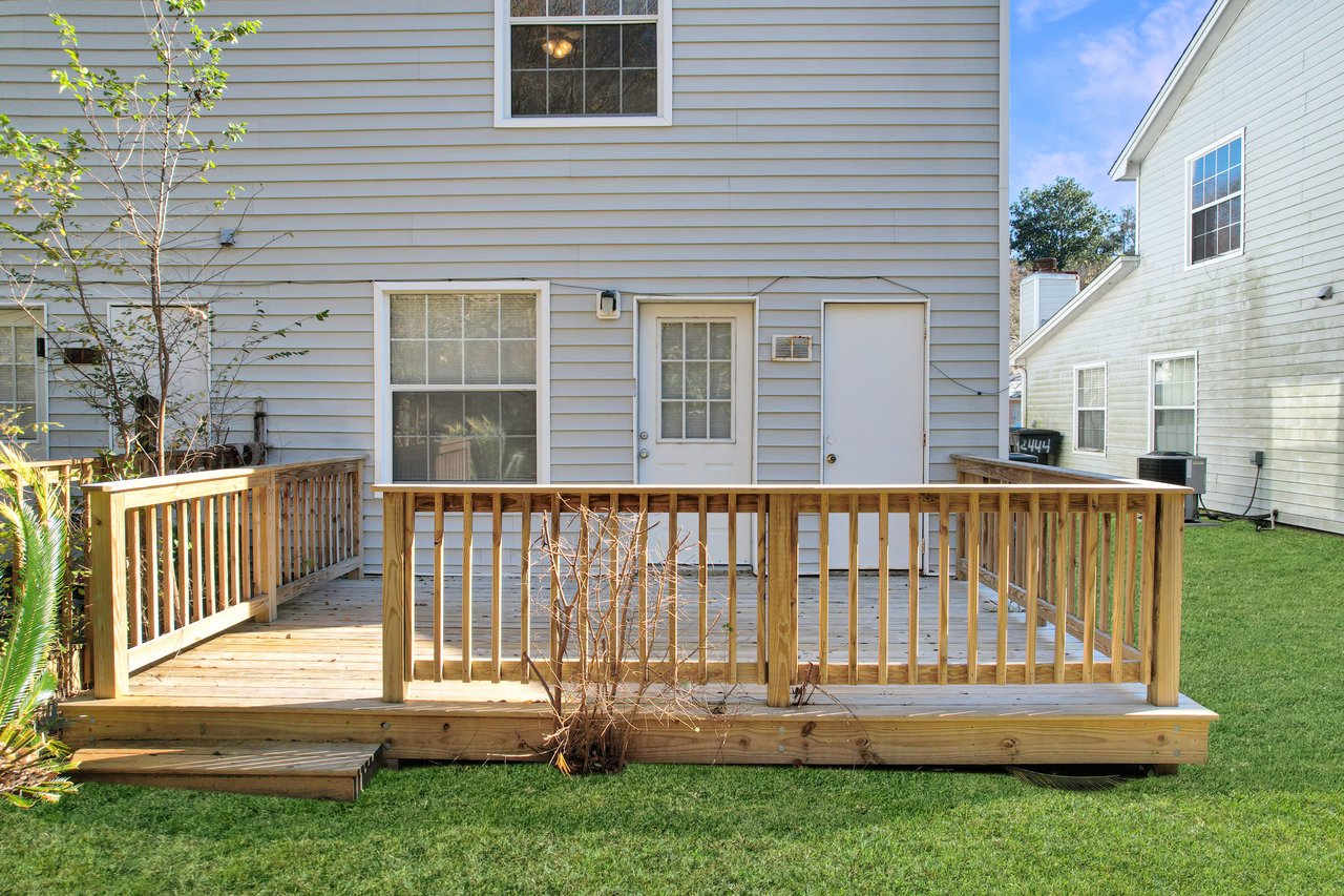 A backyard deck with light wood railings, against a gray house with two doors. A small tree is in front, and green grass surrounds the deck.