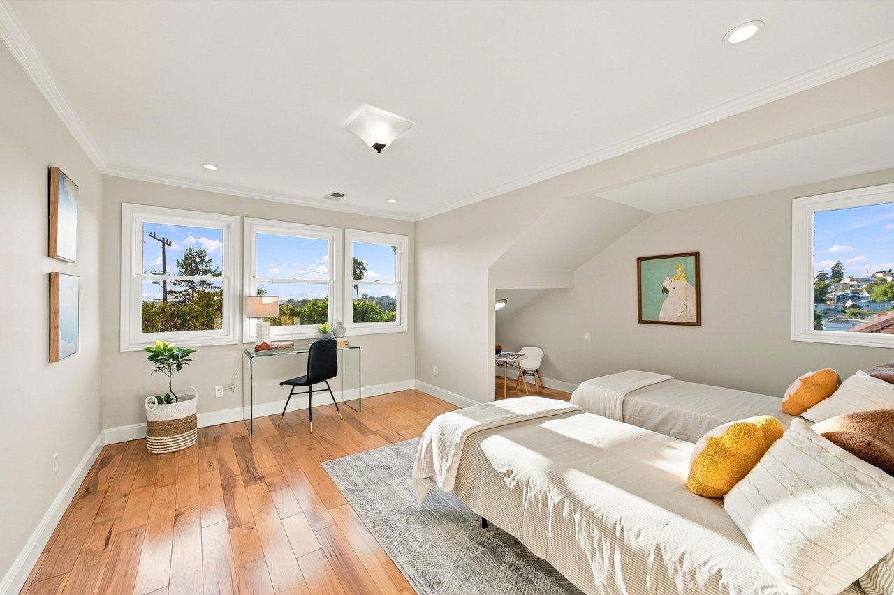 Bright bedroom with two beds, wood floors, and a desk beneath three windows.