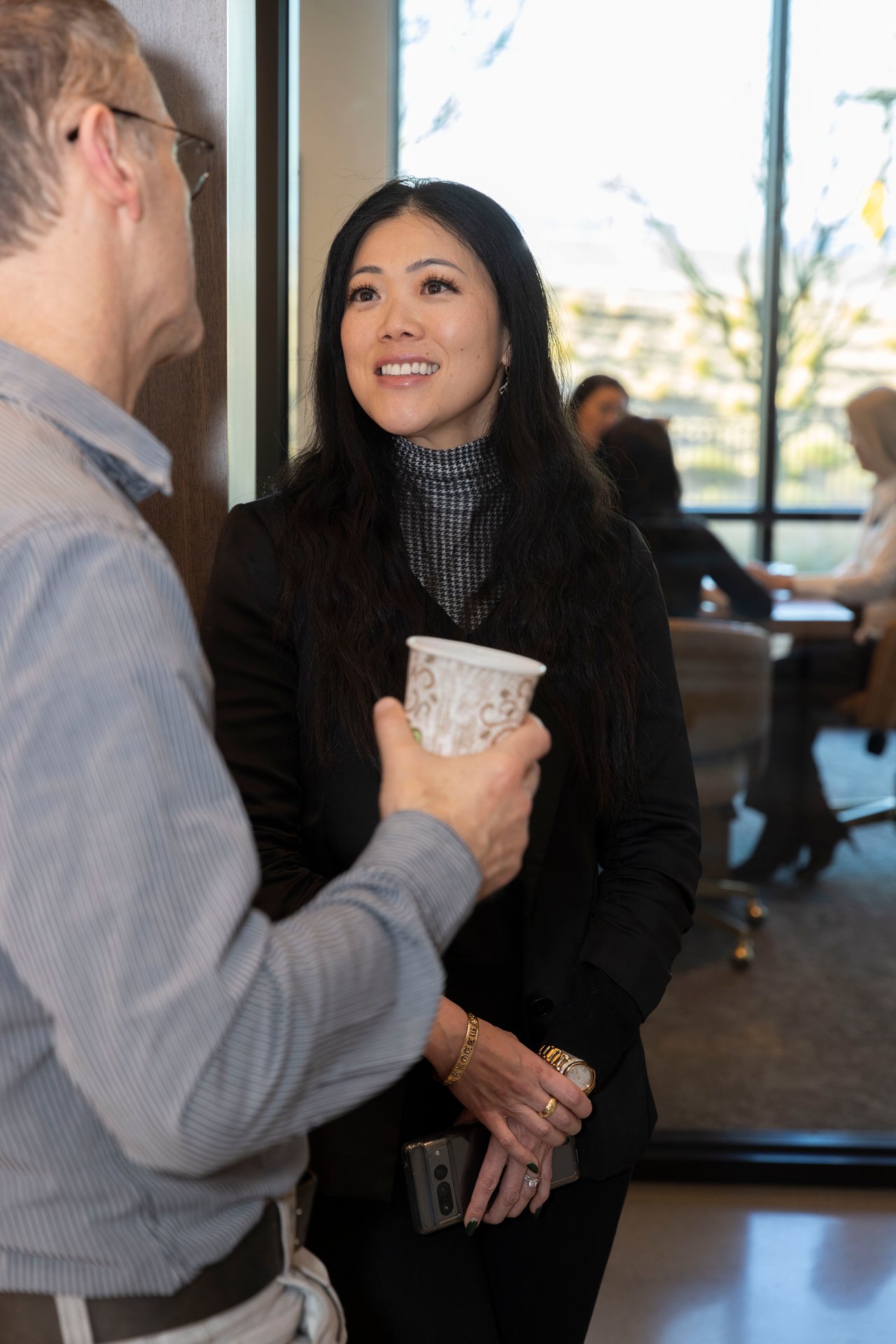 A smiling woman chats with a guest at an indoor open house, holding her phone as he offers a paper cup.