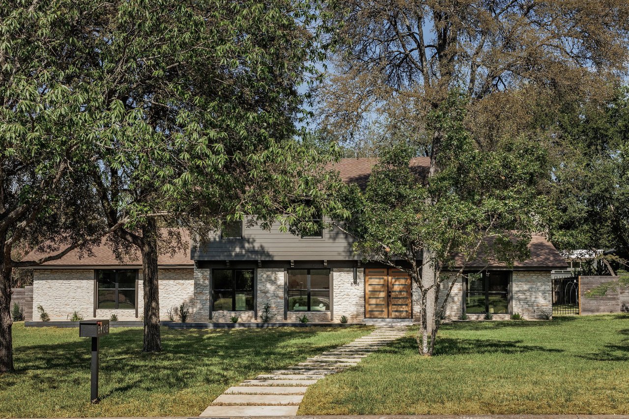 Stone-front home with double wooden doors, mature shade trees, and a stepping-stone walkway.