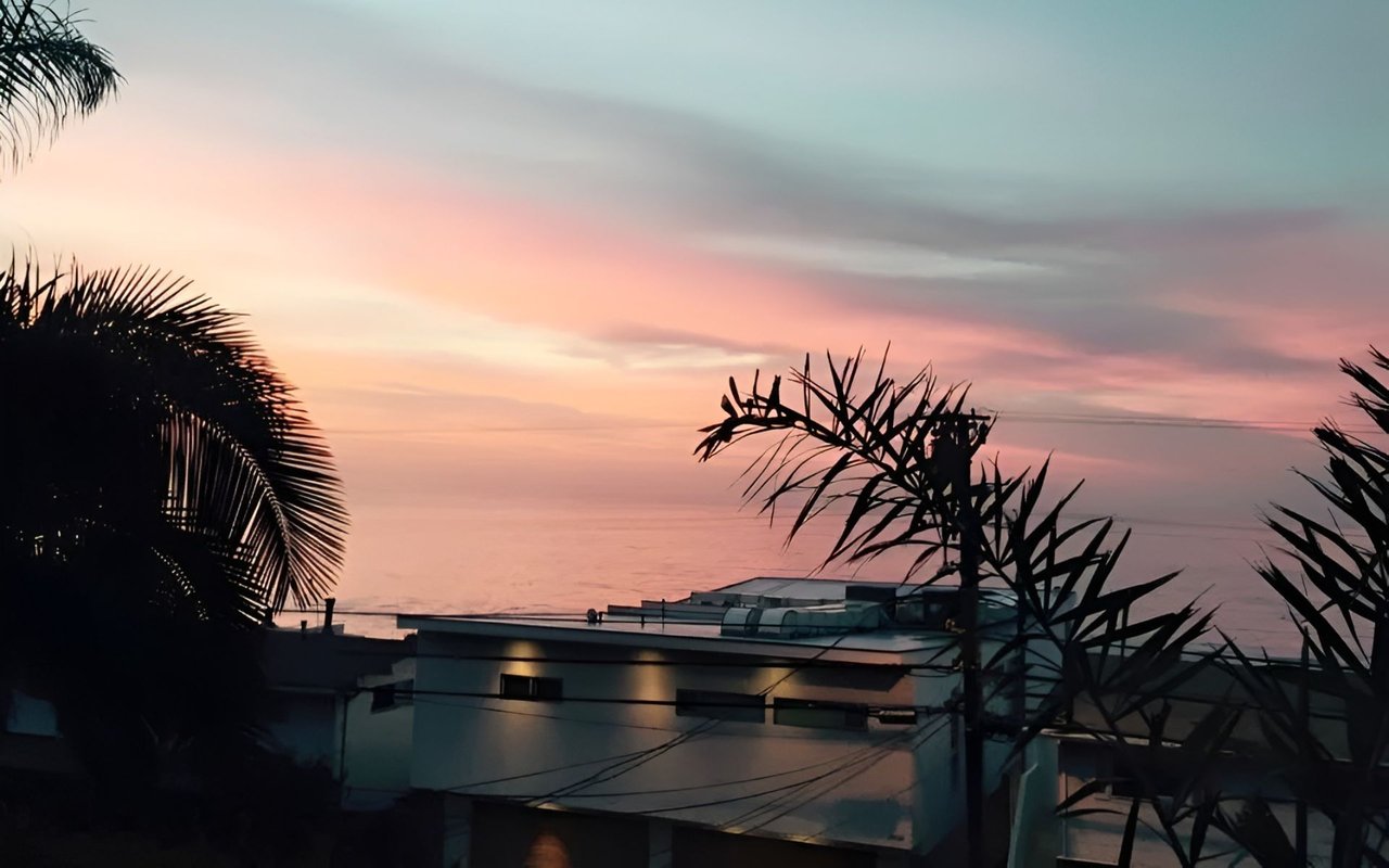 Modern beachfront house and calm ocean beneath a pastel pink morning sky, framed by tropical foliage.