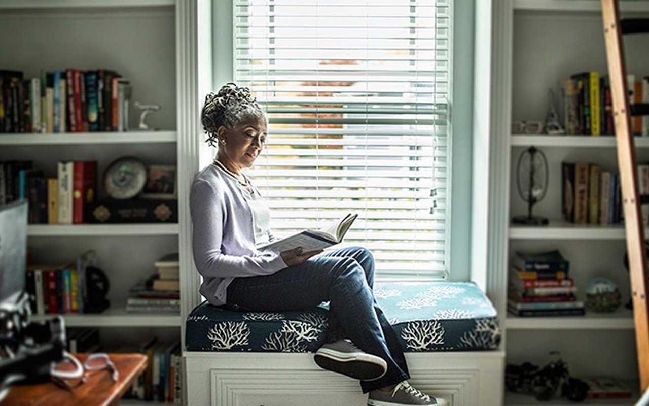 A woman reads on a cushioned window seat between built-in bookshelves.