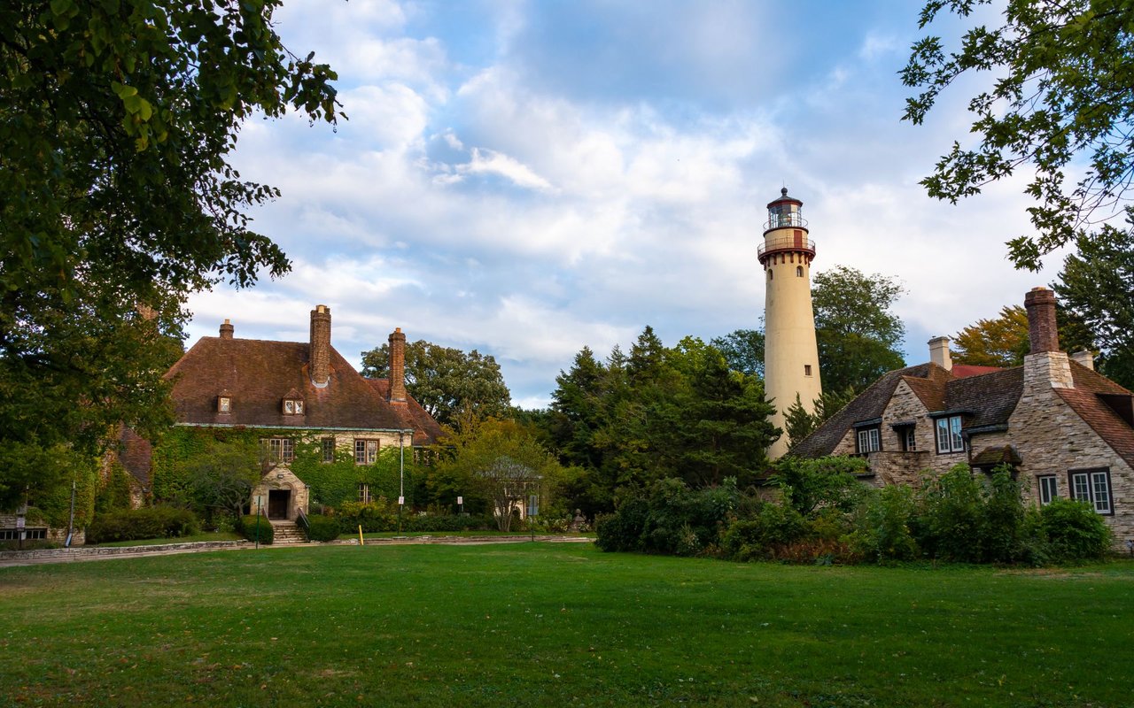 Lighthouse rising behind stone buildings and trees across a green lawn.