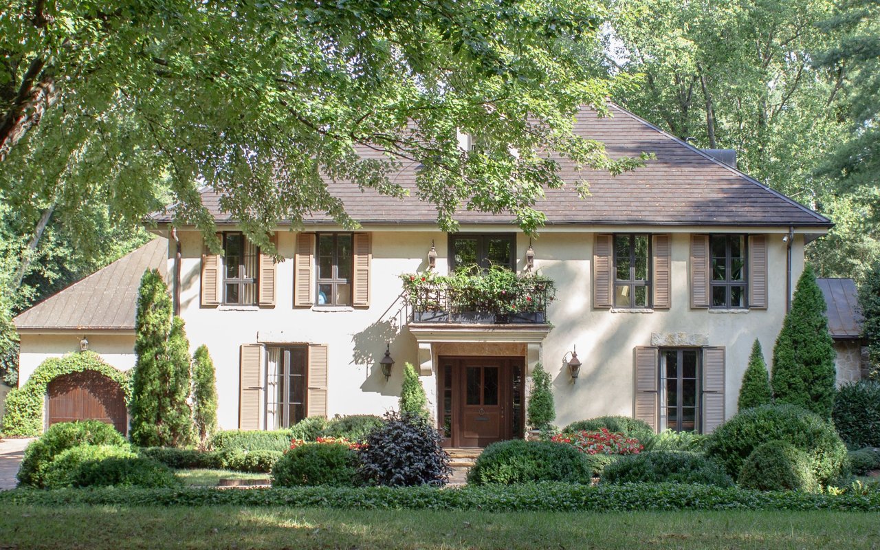 Two-story cream house with a brown roof, shuttered windows, and landscaped front garden beneath mature trees.